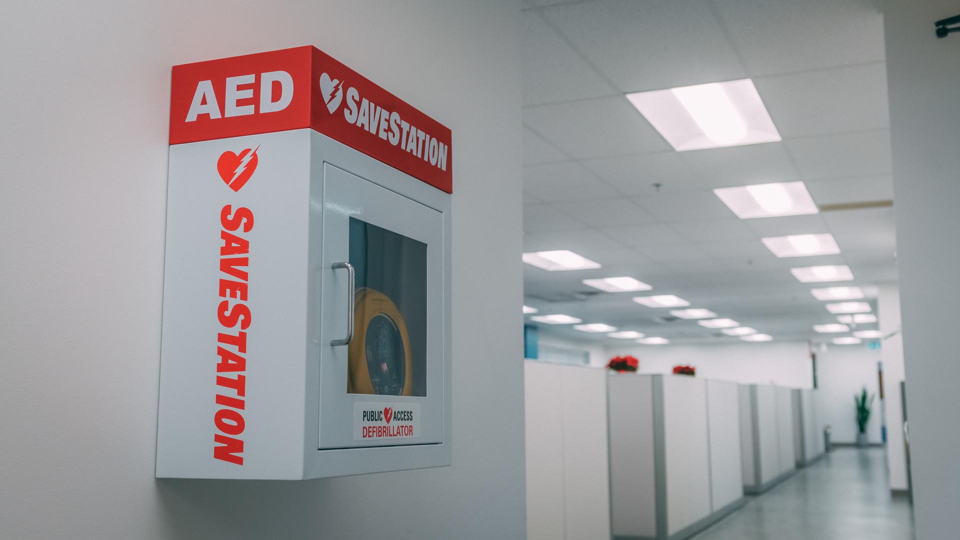 An AED (Automated External Defibrillator) cabinet mounted on a white wall in a hallway. The cabinet is white with red text 'AED' and 'SAVELATION' prominently displayed. The hallway appears to be in an office or healthcare setting with fluorescent lighting visible on the ceiling.